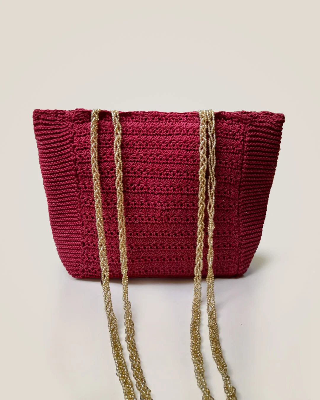 A deep red crochet tote with two long, braided gold straps.
